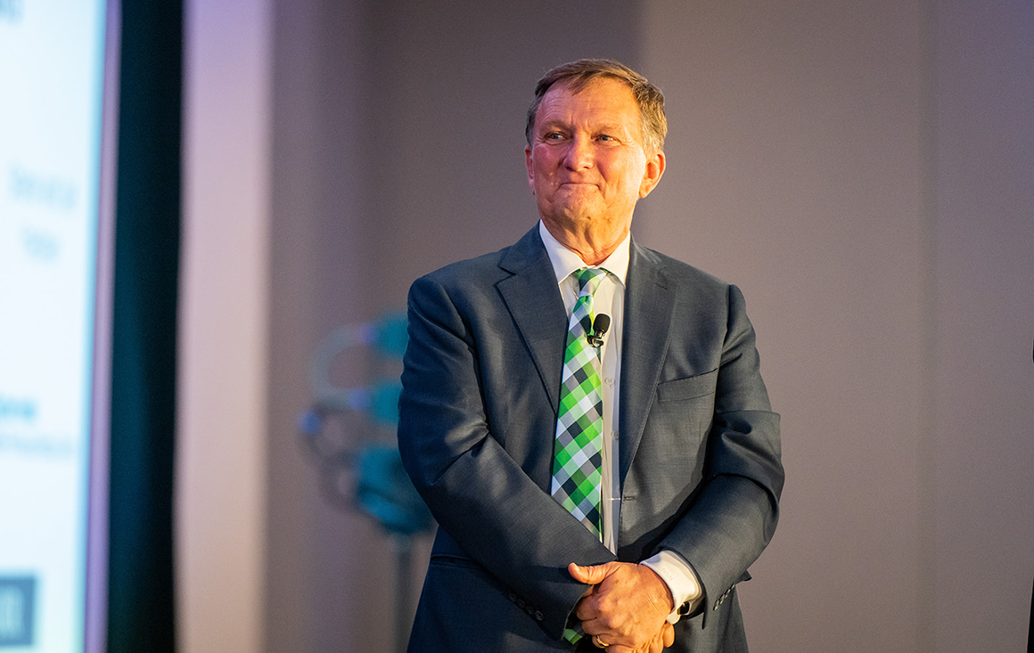 An older man in a suit stands at an event, beaming with pride.