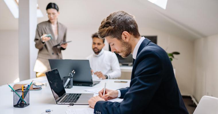 Three people work in a modern office; one man in a suit writes at a desk, another man uses a laptop, and a woman stands in the background holding a tablet. image link to article