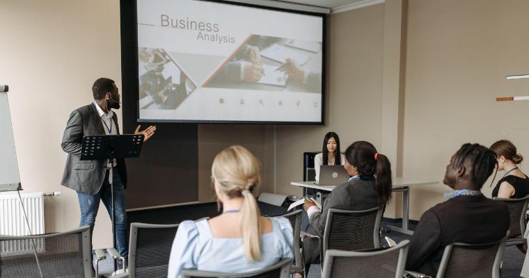 A man presents a business analysis in a conference room, gesturing towards a screen as seated attendees listen attentively. image link to article