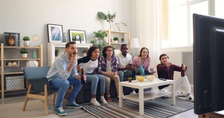 A diverse group of six friends excitedly watching TV together in a cozy living room, with a bowl of popcorn and drinks on the coffee table. image link to article