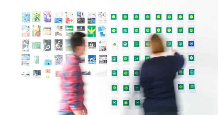 Two people interact with a wall of tennis-related images and logos, one reaching out to a logo while the other walks by. image link to article
