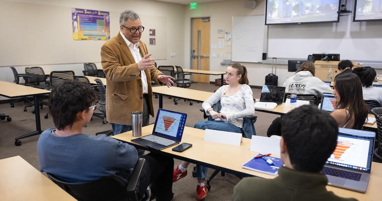 Professor Santoro speaks with students in a classroom while they work on laptops displaying charts.