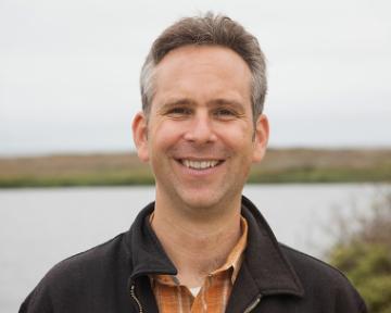 A person smiling outdoors with water and vegetation in the background.