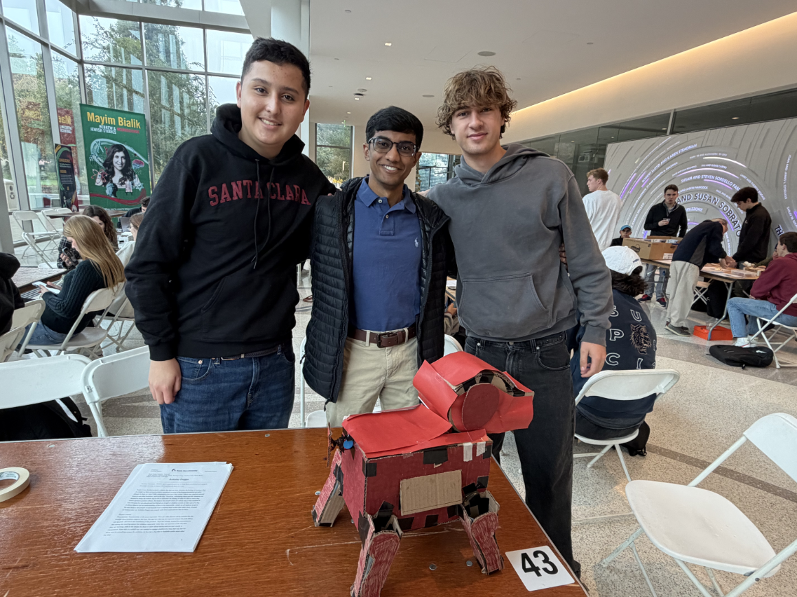 Three male students posing at a table with their red cardboard robot dog